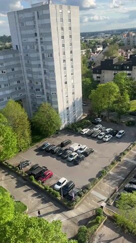 The video shows a high-angle, daytime view of a suburban neighborhood, likely taken from a tall building. Two gray, high-rise apartment buildings are prominent in the foreground, with parking lots filled with cars at their base. Beyond the apartments, there is a residential area with houses and trees. The sky is partly cloudy, with the sun shining brightly. A lens flare appears when the camera pans toward the sun.  A person is seen walking in the parking area in the beginning.