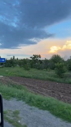 The video shows a blue road sign in Russian that says "MESTO PRIZHDENIYA YU.A. GAGARINA" and has an arrow pointing to the right. There is a pedestrian crossing sign next to it. The scene is set at dusk with a cloudy sky and some trees in the background. The camera is moving, giving a sense of traveling through the area.