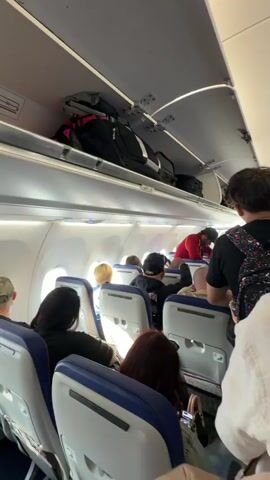 The video shows the interior of an airplane during boarding. The camera is held by a passenger looking forward down the aisle. Passengers are seated in blue and white seats on both sides of the aisle. Overhead compartments are open, containing luggage. Several passengers are visible, some standing and some seated. One passenger in the foreground is wearing a white jacket. Another passenger further down the aisle is wearing a black tank top and patterned pants. The lighting is typical for an airplane cabin.