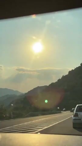 The media depicts a scenic drive on a highway through a mountainous region, likely Gornja Brova, Montenegro. The sun is prominently visible in the sky, casting a bright, warm light and creating lens flares. The time of day appears to be late afternoon or early evening, given the low angle of the sun and the golden hue of the light. The weather seems clear and pleasant, with a few scattered clouds.

The scene is dominated by a winding road with multiple lanes, suggesting it's a significant route. Several cars are visible on the road, indicating ongoing travel. The landscape is lush and green, with steep hillsides covered in dense vegetation. The road itself has white lane markings, and a guardrail is visible along the side. The perspective is from inside a vehicle, looking out through the windshield, with the dashboard visible in the foreground.

There's a distinct mood of serene travel and appreciation for the natural beauty of the surroundings. The bright sunlight and the open road evoke a sense of freedom and journey.

Visible text includes license plates on the cars. One license plate clearly reads "CA 838-TZ". The overall impression is a peaceful journey through a picturesque and natural environment.