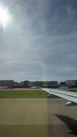 The video shows an airplane taxiing on a runway under a partly cloudy sky, with the sun shining brightly. The plane's wing is visible in the lower right corner of the frame. In the background are several airport buildings, other airplanes, and green grassy areas. The runway is concrete.
