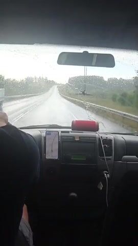 The video is taken from inside a vehicle looking forward through the windshield. It is raining heavily, with raindrops visible on the glass and the road ahead appearing wet and reflective. The vehicle is driving on a highway, with a guardrail visible on the right side of the road and trees and foliage beyond that. The dashboard of the vehicle is visible, including a smartphone mounted to the windshield displaying a navigation app. A red portable speaker sits on top of the dashboard. The steering wheel is partially visible on the left, with a person's hands on it. The headrests of two other seats are visible in the foreground, suggesting there are other occupants in the vehicle. The windshield wipers are briefly seen in motion. The overall scene depicts driving in inclement weather conditions.