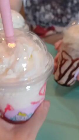 The video shows two plastic cups with domed lids and pink straws filled with creamy drinks topped with whipped cream. The cup in the foreground is held by a person's hand. The cup has a colorful unicorn-themed design on its lower portion. The other cup, slightly behind and to the right, appears to be held by another person. In the background, two people are visible. One person on the right is wearing a white t-shirt with a dark graphic design. The other person on the far left is partially visible, wearing a dark and white patterned shirt. The setting appears to be a casual indoor environment, possibly a cafe, with a teal-colored table surface. The lighting is bright, highlighting the drinks.