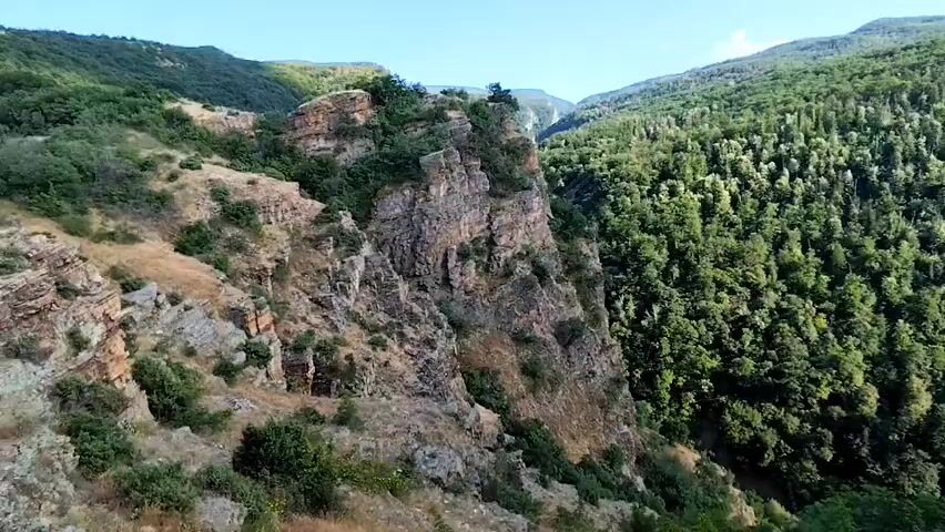 This is a landscape video of a mountain gorge in Hrabrino, Bulgaria. The scene showcases steep, rocky cliffs on the left, covered in sparse vegetation and scree. A dense, vibrant green forest dominates the right side of the gorge, with a variety of trees creating a lush canopy. In the distance, rolling mountains covered in similar forest extend towards a clear blue sky.

The video appears to be filmed during the daytime, as indicated by the bright sunlight casting shadows and illuminating the scene. The weather seems fair and clear. The perspective is from a high vantage point, looking down into the gorge and across the forested slopes. There is no visible text in the media file. The overall impression is one of natural beauty and rugged wilderness.