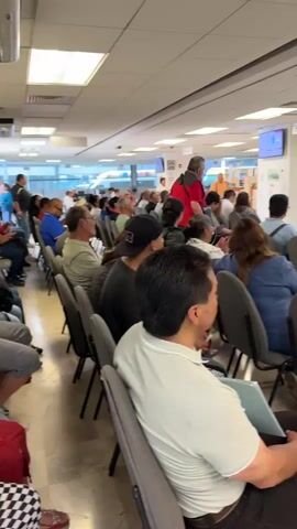 The video shows a crowded waiting area. Many people are sitting in gray chairs, facing toward a bank of teller windows. Some people are standing. One woman in a white top is speaking to some of the seated people. Other people are standing at the teller windows, speaking to the clerks. The area is lit by overhead fluorescent lights. The floor is tiled.