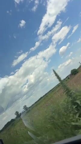 The view from a moving vehicle shows a sunny day with a blue sky dotted with fluffy white clouds. The landscape consists of a green field that turns into a patch of ploughed land, with some trees along the horizon. The metal barrier of the road is seen through the vehicle window. Faint music is playing in the background.