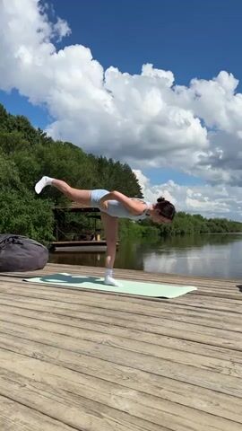 A woman performs a yoga pose on a wooden deck by a lake. She is on a mint green yoga mat, balancing on her left leg with her right leg extended backward. Her torso is tilted forward, and her arms are extended forward and outward. She is wearing a light blue sports bra and shorts, and white socks. In the background, there is a lush green forest, a body of water, and a bright blue sky with fluffy white clouds. The deck appears to be made of wooden planks.