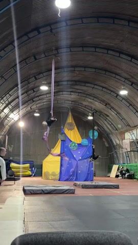 The video shows an indoor aerial performance. In the foreground, two large padded mats are visible on the floor. In the background, a vaulted ceiling with overhead lighting is apparent. A large yellow fabric structure resembling a tent is behind the performers. In front of the yellow structure is a blue fabric backdrop decorated with stars and planet-like spheres. Two aerialists are performing on silks. The aerialist on the left is on a purple silk, suspended from the ceiling. They are wearing a black leotard and perform a series of poses, including a split and a handstand. The aerialist on the right is on a black silk and is also wearing a black leotard. They are inverted, with their legs extended. Both aerialists are demonstrating flexibility and strength. The video captures the dynamic movements of the aerialists against a theatrical backdrop.