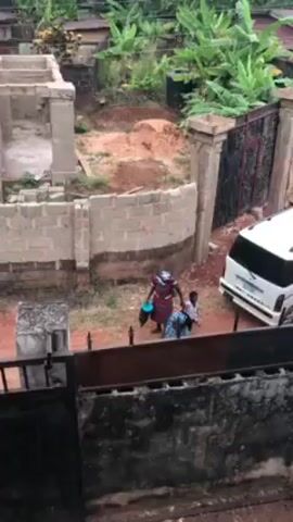 An eye-level shot shows two adults and a child outside near a building under construction. The woman is wearing a red and white patterned dress, carrying a blue bucket, and holding the child’s hand as they walk across a dirt area. The child, in a white top and black pants, carries a clear plastic bottle. An older adult is kneeling behind the child, near a blue bag. The area has a partial fence, unfinished brick walls, a white van, and various plants and trees. The video is shot through a black metal fence. The camera follows the group.