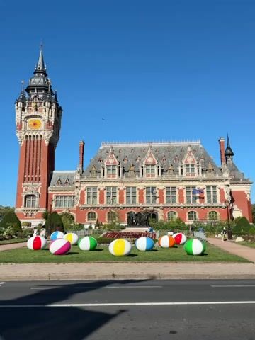 The video shows a sunny day with a clear blue sky. In the foreground, a street is visible with a gray surface and a white line marking a lane. A black car drives across the frame from left to right. In the midground, a grassy area features several large, colorful inflatable beach balls. Behind the beach balls, a grand building with a prominent red brick tower and a red roof stands under the blue sky. The building appears to be an ornate structure, possibly a town hall or similar public building, with many windows and decorative elements. In the background, the sky is clear and cloudless.