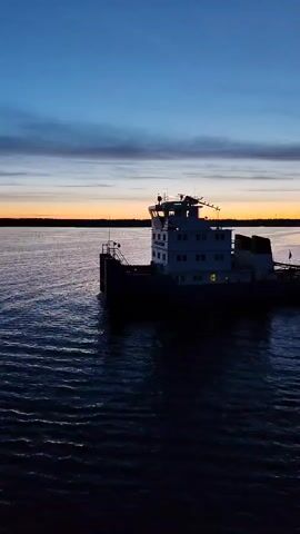 The video shows a large, white-and-black boat moving on dark water under a sky with colors of orange, purple, and blue at dusk or dawn. The boat has a multi-level structure with windows. The water is rippled, and the boat casts a reflection. The landscape in the background is dark. The video is a wide shot with the boat slightly off-center.