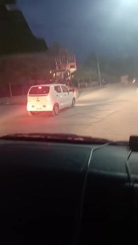 The video is shot from inside a vehicle, driving down a street at night. The street is lined with trees and streetlights. Initially, the vehicle is behind a small white car, and a large red forklift is visible on the left side of the road. The forklift appears to be driving along the street as well. A brief, loud siren-like sound is heard. The vehicle then passes the white car and the forklift, continuing down the street.