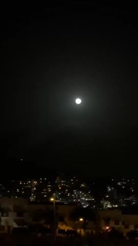 The video is a vertical shot of a night sky with a bright full moon centered. Below the moon is a cityscape with many lights visible, indicating buildings and streets. In the foreground, there are silhouettes of buildings and trees, suggesting a viewpoint from a higher elevation. The overall mood is tranquil and serene, characteristic of a quiet night. There are no people visible in the video.