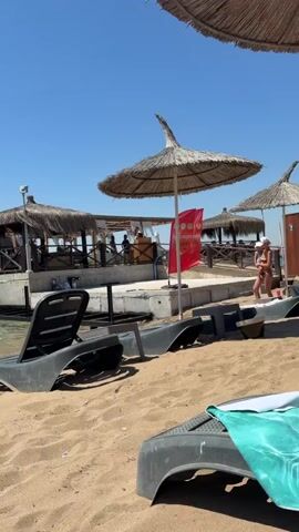 The video opens with a view of a sandy beach with several lounge chairs and umbrellas. In the background, there is a building with a thatched roof and wooden railings, appearing to be a restaurant or bar situated over the water. The water is a clear blue, and the sky is a vibrant, cloudless blue. 

As the video progresses, people are seen on the beach and in the water. Two individuals are standing near a lounge chair and an umbrella, with one appearing to be folding a towel. A young boy walks towards the water. Several other people are visible in the distance, some swimming or wading in the sea. The scene depicts a typical sunny day at a beach resort.