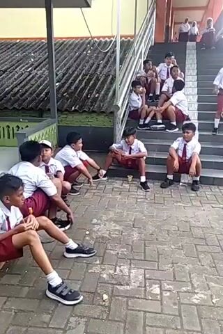 The video shows a group of young students in school uniforms sitting and standing on outdoor stairs and on a brick-paved courtyard. Some students are sitting on the steps, some are sitting on the ground, and some are standing. A few students are interacting with each other, while others are looking around. There is a building with a red roof and a staircase in the background. There are also some benches visible in the courtyard. The overall impression is a casual scene of students during a break or waiting for an activity.