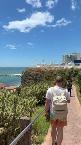 The video shows a man walking along a paved path, away from the camera, on a sunny day. He is wearing a white t-shirt, patterned shorts, and a backpack. A second person is visible further ahead on the path. The path is lined with a silver metal railing on one side and green vegetation including various cacti. To the left, there is a view of the ocean, a small building with a red-tiled roof, and a breakwater. To the right, on a cliff, there is a white structure with decorative lights and, in the background, a tall white hotel building. The sky is blue with scattered white clouds.