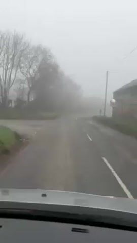 The video shows a view from inside a car driving on a narrow, winding road in heavy fog. The visibility is severely limited, with trees and a building barely visible through the fog. The road is wet and has a solid white line marking the right edge. There is a fork in the road ahead. The sky is overcast and gray. No people are visible.