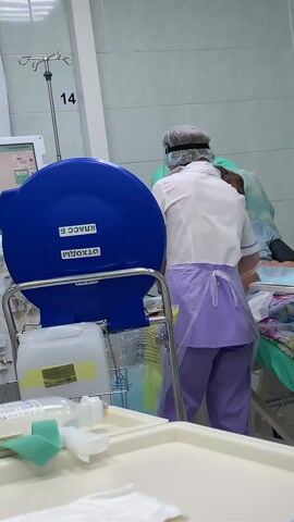 The scene takes place in a medical setting, possibly a hospital room, identifiable by the equipment and attire of the individuals present. A medical worker in a white shirt, purple pants, a clear plastic apron, and a surgical cap with a face shield tends to a patient lying on a hospital bed. The patient is an older man with short hair, wearing a light blue shirt. The medical worker is positioned behind him and appears to be attending to his left arm, which is covered with a blue medical glove.  A blue container labeled "отходы класс Б" is visible in the foreground, suggesting it is a biohazard waste disposal unit. Other medical equipment is scattered around, including an IV stand with the number "14" marked on it, a small white tray with various medical supplies, and an electronic monitor displaying medical data. The walls of the room are a light color, possibly tile or painted with a hygienic coating.