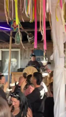 The video captures a vibrant party scene, focusing on a DJ performing for a crowd of people. The DJ, wearing a dark cap and shirt, stands behind a set of equipment, actively manipulating controls and engaging with the audience. A large decorative circle with a floral pattern is placed in front of the equipment, adding to the visual appeal. The crowd consists of people of diverse ages, dressed in various casual outfits, moving and dancing to the music. The environment features a wooden structure with a roof decorated with colorful streamers and fabric, creating a festive atmosphere. Lighting fixtures are visible, suggesting the event may take place during the day, enhanced by artificial lighting. The overall mood is lively and energetic, indicative of a successful party or gathering.