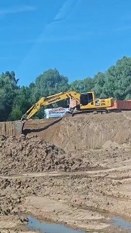 A yellow Komatsu excavator is digging a large pile of dirt on a construction site. The excavator is positioned on top of a dirt mound, and its arm is extended to scoop dirt. A truck is parked next to the excavator, and it appears to be receiving the dirt. The background of the scene is a blue sky and trees. The video is shot from a distance, and the image is slightly blurry.