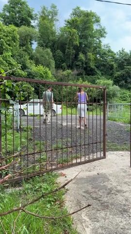 Two individuals, a man and a woman, walk away from the camera through an open metal gate. The man is wearing a green t-shirt and beige pants, while the woman is wearing a purple tank top and a white skirt. A white car is parked behind them to the left. The ground appears to be covered in gravel.

The scene is set outdoors, with lush green trees and foliage forming a backdrop behind the gate. The overall impression is of a casual, possibly rural, setting. The sky is visible and appears overcast, suggesting a cloudy day. The time of day is indeterminate but likely daytime given the ambient light. There is no visible text in the media file. The activity is simply walking, with no clear indication of purpose or emotion. The location context of Shepsi, Russia, might suggest a natural environment, possibly near a residential area or a park, given the presence of a car and a gate.