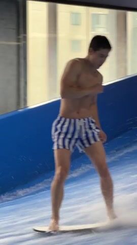 A man is surfing on an indoor wave machine. He is wearing blue and white striped swim trunks and is standing on a surfboard. He is in the process of falling off the board and into the water. The wave machine is in a large indoor space with a window that looks out onto a city skyline. There are colorful lights in the background.