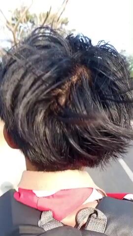 The video shows a close-up view of the back of a person's head and neck. The person has dark, somewhat messy hair. They are wearing a backpack with grey straps. Around their neck, they have a red scarf or bandana over a light-colored collar. The person appears to be walking or moving, as evidenced by the slight motion blur and the way their hair moves. The environment seems to be outdoors, possibly on a path or road, with some blurred foliage visible in the background. The lighting suggests it is daytime.