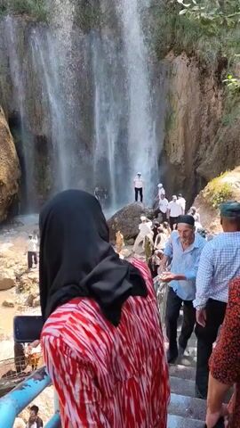 A group of people are gathered near a waterfall. The waterfall cascades down a rocky cliff face. Some people are standing on rocks near the base of the waterfall, while others are walking on a path. The area is lush with greenery. Some individuals are wearing headscarves. One man in the foreground is wearing a blue shirt and a white cap. Another person in the foreground is wearing a red and white striped garment and a black headscarf. The water appears to be muddy.