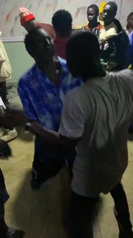 The video shows a group of young people dancing in what appears to be a dimly lit indoor space. In the foreground, a person in a white t-shirt is captured from behind, facing away from the camera and towards other individuals. To the left and slightly in front of the person in white, another individual in a blue and white patterned shirt is visible, also dancing. Several other people are in the background, some watching and others participating in the dancing. One person in the background is wearing a red and white hat. The walls in the background are decorated with a colorful, abstract pattern. The floor is a light-colored tile. The lighting is low, creating shadows and highlighting the movement of the dancers.