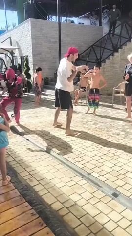 A group of people are gathered outdoors on a paved area. In the foreground, a person in a Deadpool costume is dancing and performing acrobatic moves. To their left, a man in a white t-shirt and black shorts and a red baseball cap is also dancing. Behind them, a young boy in striped swim trunks stands with his hands on his hips. Other individuals are visible in the background, some standing and some seated. A set of outdoor stairs with a black railing is to the right of the frame, with people on the stairs and at the top. The environment appears to be a recreational area, possibly near a pool, with some greenery visible. The lighting suggests it is daytime.