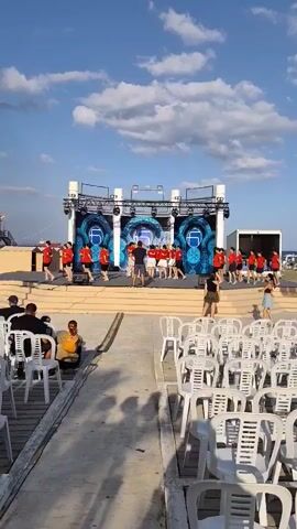 A group of young people in red shirts are performing a dance routine on an outdoor stage. The stage has a large screen behind the performers displaying a logo. White chairs are set up in front of the stage for an audience, with some people already seated. The sky is blue with scattered white clouds, suggesting it is daytime. The ground appears to be a wooden boardwalk or platform. There is a raised walkway leading to the stage area.