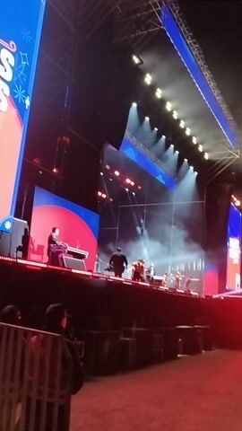 The video captures an elevated view of a vibrant stage performance taking place at night. Multiple performers are visible on a large stage, illuminated by numerous spotlights and red stage lights. Some performers appear to be dancing or moving to music, while others stand with what looks like musical instruments, including a drum set on the right side of the stage. Fog or smoke effects emanate from the stage, enhancing the atmosphere.

The stage features a dark main area with distinct blue and red sections on the stage floor, along with large display screens showing dynamic graphics. One screen on the far left shows a blue background with white star-like shapes and partially legible stylized text, possibly "sa" or "as." The stage structure includes extensive trussing for lighting and screens. The overall setting suggests a concert or large-scale entertainment event. The location is Maipú, Chile.