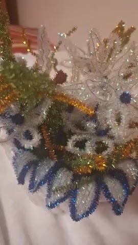 The video displays a close-up, slightly blurred view of a decorative object, possibly a wreath or a craft item, made of tinsel and pipe cleaners. The object is adorned with various colors, including green, gold, white, and blue, arranged in floral or feather-like patterns. Some parts of the decoration sparkle due to the reflective material. The background is a plain, light-colored surface, likely fabric, which appears soft and possibly textured. The lighting is warm and creates highlights and shadows on the object, suggesting an indoor setting with artificial light. The video seems to be a static shot, focusing on the intricate details and shimmering textures of the decoration. There are no people or visible actions taking place within the frame.