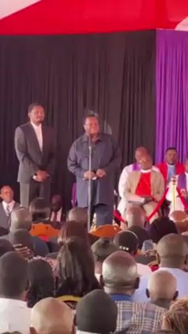 The video shows two men standing at a microphone speaking to a crowd, presumably at a formal event. The man at the microphone is wearing a dark blue dashiki-style outfit. To his left stands a man in a dark suit with a light-colored shirt and no tie, hands clasped in front of him. Behind them are seated religious figures in robes. The audience consists of individuals of varying ages and genders, mostly seated, facing the speakers. The setting appears to be under a tent, decorated with red, black, and purple fabrics.