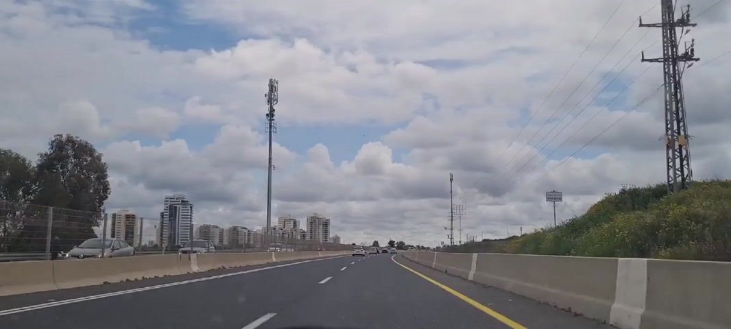 The video shows a highway with multiple lanes in both directions. A yellow line separates the rightmost lane from the shoulder. Concrete barriers line both sides of the road. On the left side, there are several modern-looking high-rise buildings in the distance. On the right side, there is a grassy area with yellow wildflowers. In the sky, there are numerous puffy white clouds against a blue backdrop. Several cell phone towers are visible along the side of the road. Cars are driving in both directions. A sign is visible to the right.