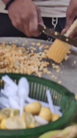 The video shows a close-up of someone preparing corn. In the foreground, a basket filled with lemons and plastic spoons is visible. The person is using a knife to cut corn kernels off a cob, which are falling into a large, shallow metal bowl. Scattered around the bowl are discarded corn husks and kernels. In the background, a glimpse of a motorcycle is visible. The scene appears to be outdoors, likely at a street food stall, with a simple brick structure containing a fire for cooking in the lower right corner. Later in the video, several small paper bowls filled with the prepared corn, each with a plastic spoon, are placed on a flat metal surface that is heated by the fire below.