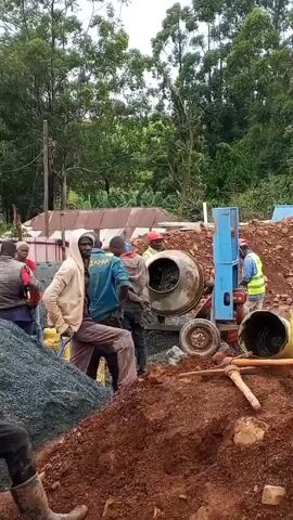The video shows a construction site with several people working. A cement mixer is in operation, surrounded by workers, some wearing safety vests and hard hats. A pile of gravel and a mound of reddish dirt are present. Some workers are carrying yellow buckets. The background features a line of trees and a glimpse of a building with a corrugated metal roof. The sky is overcast.