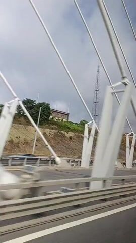 A view from a moving vehicle on a bridge shows a large structure of cables and supports. Beyond the bridge, a hill with vegetation and a building is visible, topped by a tall communication tower. The sky is cloudy. The road surface appears to be asphalt with white lane markings.