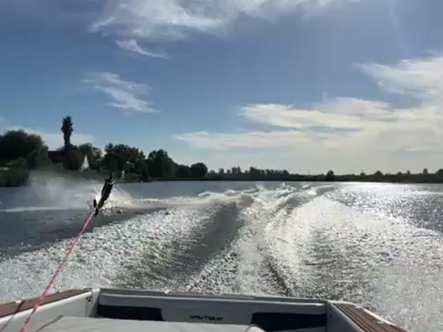 The video shows a waterskier being pulled behind a Nautique boat on a body of water, possibly a lake or river, on a sunny day. The waterskier, who appears to be wearing dark clothing, is visible in the center of the frame, creating a spray of water as they move. The wake of the boat is prominent, forming a V-shaped pattern behind it. The shoreline is visible in the background, featuring trees and some foliage. The sky is blue with scattered clouds. The sun is bright, creating a lens flare effect in the video.