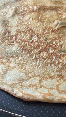 The video shows a close-up view of a crepe cooking in a pan. The crepe is golden brown with a lacy pattern on its surface, indicating it is being cooked evenly. The pan appears to be non-stick, with a dark, textured surface. The camera angle is overhead, focusing solely on the crepe and the pan. There are no people or other objects visible in the frame. The lighting is warm and natural, suggesting the video was filmed in a kitchen environment. The crepe is slowly moving and bubbling as it cooks.
