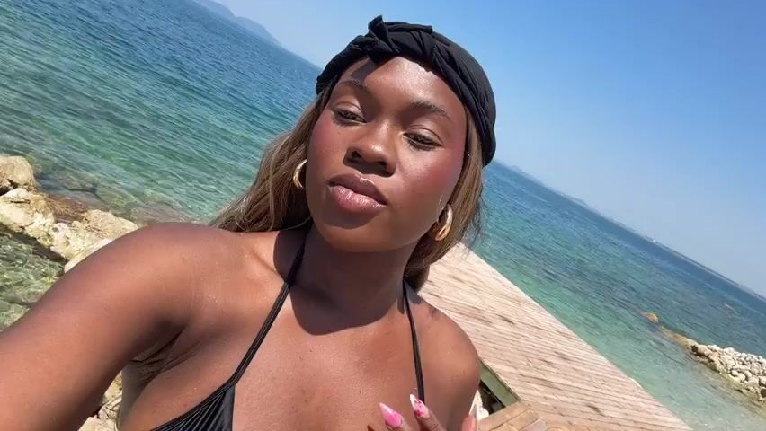 A young Black woman with blonde hair and a black headscarf is taking a selfie at a beach. She is wearing a black bikini. The ocean is to her right, with a wooden pier extending into it. In the background, there is a kayak with people in it, rocks, and mountains. The sky is clear and blue.