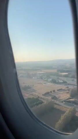 This is a vertical, close-up, outdoor shot filmed from an airplane window. The view looks out at a landscape of brown and green fields, a road, and some buildings. The sky is a clear blue with a hazy horizon. The airplane window frame is visible in the foreground.