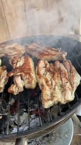 A close-up, straight-on shot shows several pieces of meat and a slice of onion grilling on a charcoal grill. Smoke rises from the meat as it cooks over the open flames. The meat is spaced out on the grill, and grill marks are visible on each piece. The grill is black and the coals are glowing orange. A wall of wooden planks is visible in the background. The video is slightly out of focus in the initial seconds but quickly sharpens.