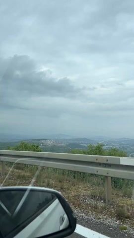 The video captures a panoramic view from the passenger side of a moving vehicle, looking out over a landscape under an overcast sky. In the foreground, the bottom left corner features a prominent reflection in a white car's side mirror, showing parts of the car's interior. A sturdy, light-grey metal guardrail runs horizontally across the lower third of the frame, separating the road from the natural terrain below.

Beyond the guardrail, the immediate hillside is covered with dry grass, small plants, and rocks, indicating a somewhat rugged, uncultivated area. Further down the slope, lush green trees and dense foliage dominate the mid-ground, stretching towards a distant valley.

In the valley, several clusters of buildings and what appears to be a town or industrial complex are visible. A hazy mountain range forms the backdrop on the horizon. The sky is heavily clouded with various shades of grey, suggesting an overcast day with diffused lighting; there is no direct sunlight.

The slight movement of the scenery, particularly the passing trees in the foreground, indicates that the vehicle is driving along an elevated road, likely a mountain pass or a highway offering an elevated perspective of the Gornji Kokoti region in Montenegro.

A pop song with a rhythmic beat and vocals is playing in the background, adding an auditory element to the scene. There is no visible text in the video frame.