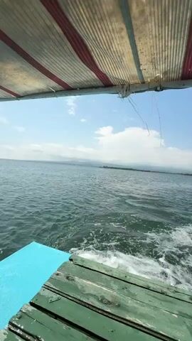 The video begins by showing the wake of a boat as it moves through the water. The camera is positioned at the rear of the boat, looking out over the water. The boat has a striped awning overhead. The water is calm and blue, with some clouds in the sky. In the distance, there are some trees and buildings on the shore. The video then pans to the left, showing more of the shoreline and some docks. The boat continues to move through the water, and the wake becomes more pronounced. The video ends with a view of the wake and the sky. There are two people's feet visible at the bottom of the frame, suggesting they are seated on the boat.