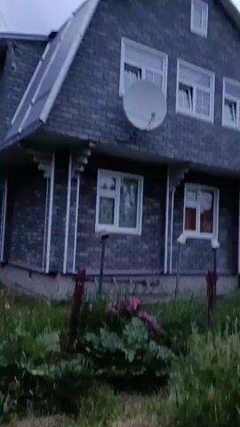 The video begins by showing a dark gray wooden house with a pitched roof. A white satellite dish is mounted on the roof. The house has several windows with white frames. In front of the house, there are green plants and a few pink flowers.

The camera then pans to the left, showing a tall, gray utility pole. Behind the pole, there are trees and bushes. The camera then pans back towards the house, showing more of the front yard.

Next, the video shows a path made of gravel and dirt. A dog on a leash is walking down the path. The dog is light brown and white. There are trees and bushes on either side of the path. In the background, there is a utility pole with wires.

The video ends by showing the dog continuing to walk down the path. The sky is overcast and the light is dim, suggesting it is either early morning or late evening.