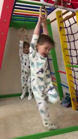 Two young children, both wearing patterned, long-sleeved pajamas, are playing on an indoor play structure. The child in the foreground is suspended from a pair of wooden gymnastic rings, swinging slightly and looking at the camera with a smile. The child in the background is standing on the ground, reaching up to touch a similar pair of rings. The play structure is colorful, with sections painted red, green, and yellow, and includes a climbing net on one side. The floor is carpeted.