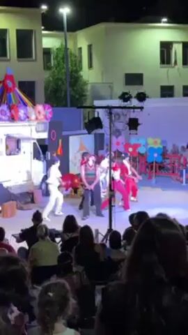 A nighttime outdoor performance is captured in a video. Five performers, dressed in clown-like costumes, dance on a stage bathed in bright purple light. In the audience, children and adults alike watch the performance. The backdrop includes a decorated van, stage props, and a building lit by streetlights. The performers' costumes vary: some in white with red accents, others in red suits, and one in a vest and pants. They move in unison, performing synchronized dance steps. The audience sits or stands, their silhouettes outlined against the light. The video is shot from the crowd, providing a viewer's perspective of the theatrical event.