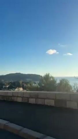 The camera films a landscape with a sea and a coastline, framed by a stone wall in the foreground.  The sky is a clear blue with a few scattered clouds. A pedestrian crossing sign briefly appears as the camera pans. The sun shines brightly, creating a glare on the water and silhouetting the land in the distance. There is a small boat on the water.