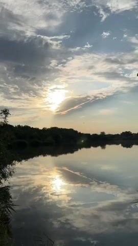 The video displays a serene natural landscape. A tranquil body of water occupies the foreground, reflecting the sky and surrounding vegetation. On the horizon, a line of dense, dark green trees forms a silhouette against the sky. The sky itself is a canvas of soft, wispy clouds, with patches of bright sunlight breaking through, suggesting either sunrise or sunset. The overall impression is one of peacefulness and natural beauty. There are no people or human activity visible.
